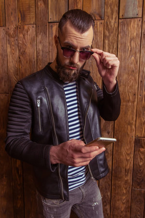 Stylish man with beard in leather jacket and sunglasses using a mobile phone, standing on a wooden backgroundの写真素材
