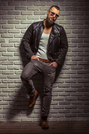 Attractive stylish man with beard in leather jacket and sunglasses looking at camera, standing against brick wallの写真素材