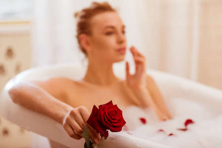 Beautiful young blonde woman enjoying pleasant bath with foam, holding a rose, focused on hand with roseの写真素材