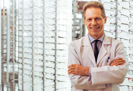 Handsome middle aged ophthalmologist looking at camera and smiling while standing in his officeの写真素材