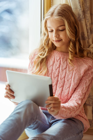 Lovely teenage girl using a tablet and smiling while sitting on the window sill in the room
