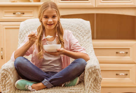 Lovely teenage girl holding a bowlful and a spoon, looking at camera and smiling while sitting on the armchairの写真素材