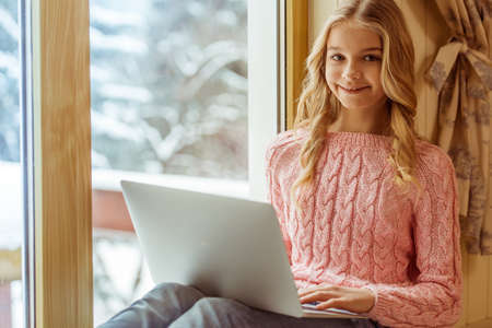 Lovely teenage girl using a laptop, looking at camera and smiling while sitting on the window sill in the roomの写真素材