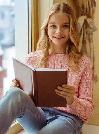 Lovely teenage girl reading a book, looking at camera and smiling while sitting on the window sillの写真素材