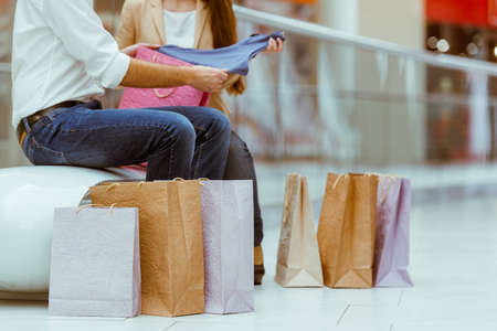 Happy beautiful young couple looking upon their purchase and smiling while sitting on a bench in mall after doing shopping, croppedの写真素材