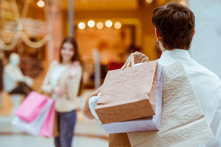 Back view of handsome man meeting his beautiful woman in mall. Both holding shopping bagsの写真素材