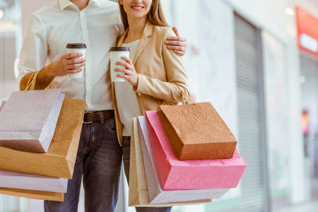 Happy beautiful young couple holding shopping bags and cups, looking upon showcase and smiling while standing in mallの写真素材