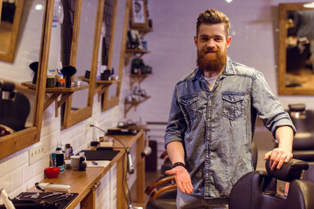 Handsome young bearded barber pointing on a chair, looking at camera and smiling while standing in...