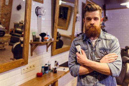Handsome young bearded barber holding a hair trimmer and looking at camera while standing in the...
