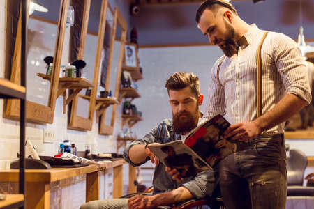 Attractive muscled bearded barber and handsome young customer reading a magazine and choosing haircut at the barber shopの写真素材