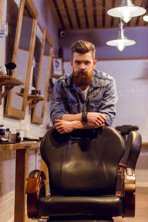 Handsome young bearded barber leaning on a chair and looking at camera while standing in the barber shopの写真素材