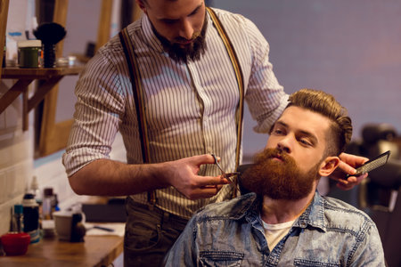 Attractive muscled bearded barber cutting handsome young customer beard at the barber shop