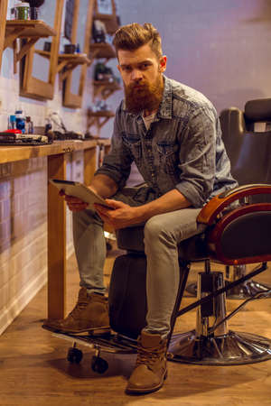 Handsome young bearded man using a tablet and looking at camera while sitting on a chair at the barber shopの写真素材