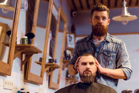 Handsome young bearded barber styling attractive bearded customer hair at the barber shopの写真素材