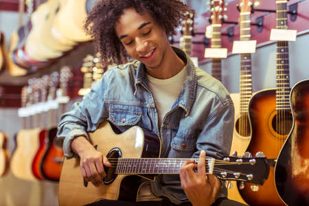 Handsome young Afro-American man in jeans jacket playing a guitar and smiling while sitting in a musical shopの写真素材
