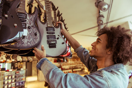 Young Afro-American man in jeans jacket smiling while choosing an electric guitar in a musical shop