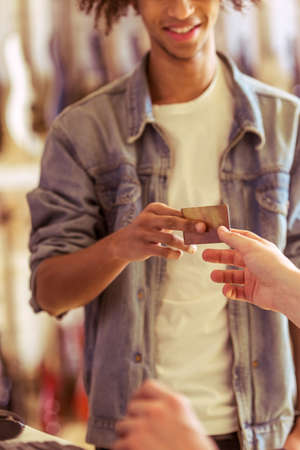 Handsome young Afro-American man in jeans jacket smiling while paying with a credit card in a musical shop, croppedの写真素材