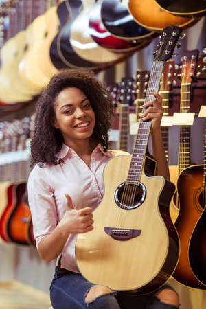 Beautiful Afro-American girl holding a guitar, showing OK sign and smiling while sitting in a musical shopの写真素材