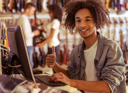 Handsome young Afro-American man in jeans jacket using a computer, showing OK sign and smiling while standing in a musical shopの写真素材