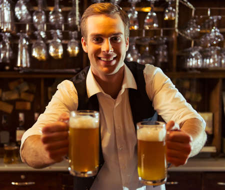 Handsome bartender is smiling, looking at camera and holding glasses of beer while standing at bar counter in pubの写真素材
