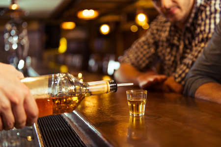 Young men in casual clothes is sitting at bar counter in pub, a bartender is pouring alcoholic beverage into glass, close-upの写真素材