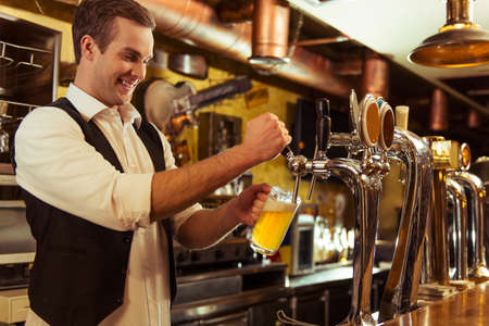 Handsome bartender is smiling and filling a glass with beer while standing at bar counter in pubの写真素材