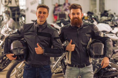 Handsome young bearded and blond men in black leather jackets are holding helmets, showing Ok sign...