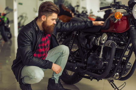 Handsome young bearded man in black leather jacket is examining his motorbikeの写真素材