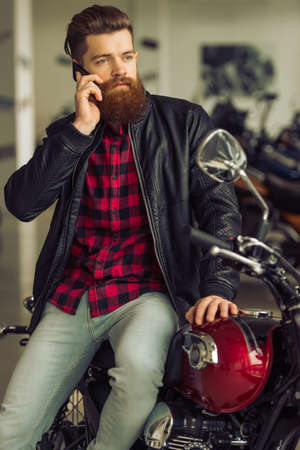 Handsome young bearded man in black leather jacket is talking on the mobile phone and looking away while sitting on motorbikeの写真素材