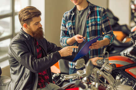 Handsome young bearded man in black leather jacket and attractive blond man are talking and smiling while examining motorbikes, croppedの写真素材