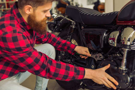Handsome young bearded man in casual clothes is repairing a motorbikeの写真素材