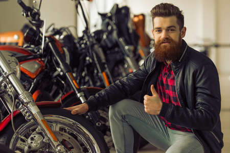 Handsome young bearded man in black leather jacket is showing Ok sign and smiling while examining motorbikesの写真素材