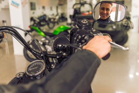 Attractive young blond man in black leather jacket is looking at his reflexion in motorbike mirror...