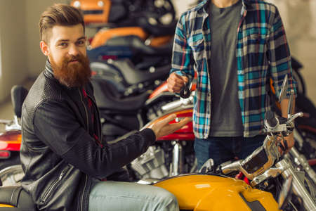 Handsome young bearded man in black leather jacket is looking at camera while buying a motorbike, attractive blond man is giving keysの写真素材