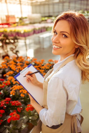 Beautiful young woman in apron is making notes about plants, looking at camera and smiling while standing in orangeryの写真素材