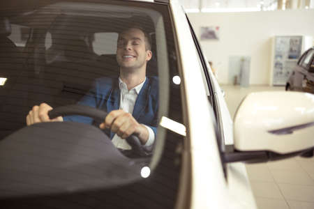 Handsome young businessman in classic blue suit is smiling and imitating ride while sitting in car in a motor showの写真素材
