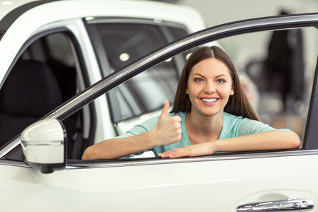Beautiful young woman in casual clothes is smiling and showing Ok sign while leaning on a new car in...