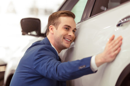Handsome young businessman in classic blue suit is smiling while examining car in a motor showの写真素材
