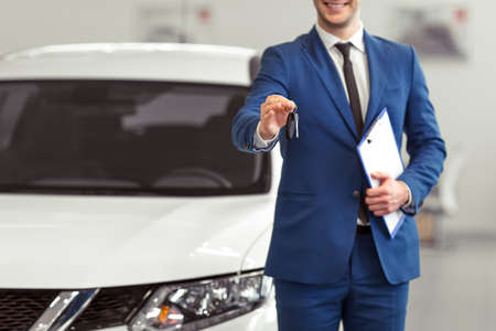 Handsome young man in classic blue suit is smiling, looking at camera and offering car keys while presenting car in a motor show, croppedの写真素材