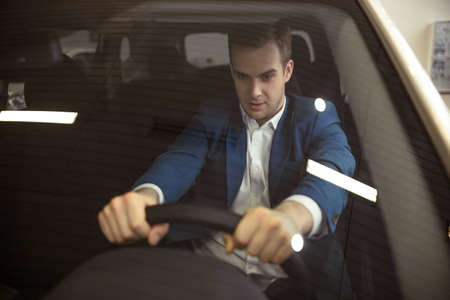 Handsome young businessman in classic blue suit is imitating ride while sitting in a new car in a motor showの写真素材