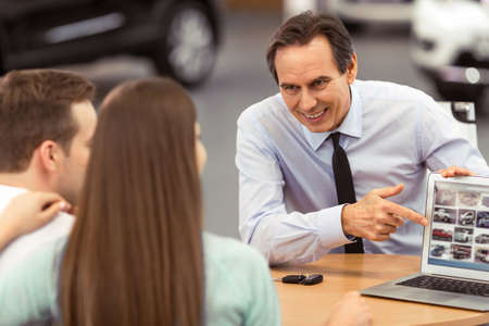 Young couple is buying a car. Handsome middle aged worker of motor show is smiling and showing cars using a laptopの写真素材