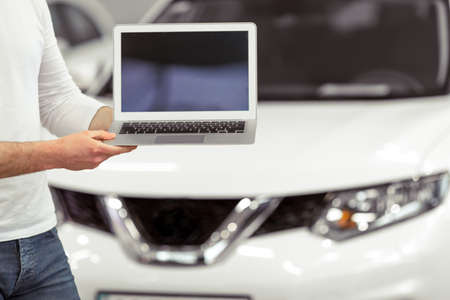 Handsome young man is holding a laptop while standing in a motor show, close-up. Modern white car in the backgroundの写真素材