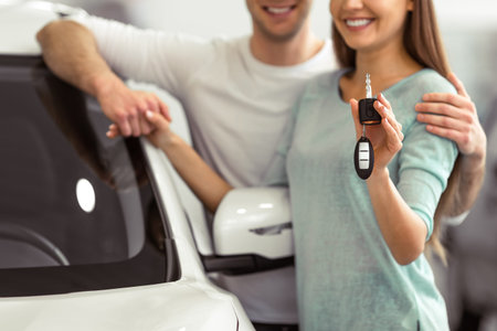 Beautiful young couple is smiling and looking at camera while leaning on their new car in a motor show. Woman is holding car keys, close-upの写真素材