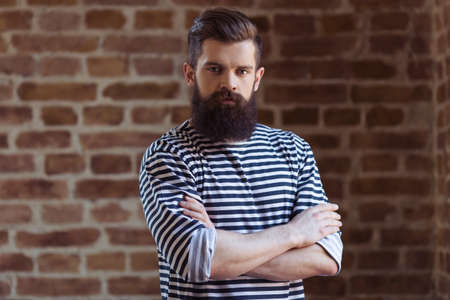 Stylish young bearded man in striped vest is looking at camera, standing against brick wallの写真素材
