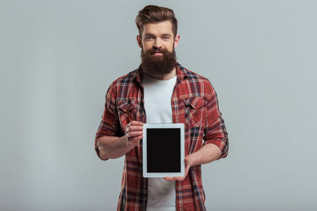 Handsome young bearded man in casual clothes is showing a tablet and smiling, on a gray backgroundの写真素材