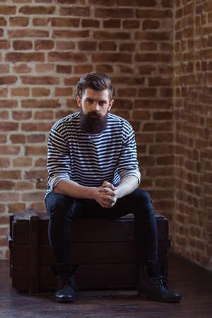 Stylish young bearded man in striped vest is looking at camera, sitting against brick wallの写真素材