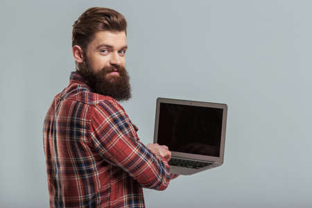 Handsome young bearded man in casual clothes is using a laptop and smiling while standing turned in a gray backgroundの写真素材
