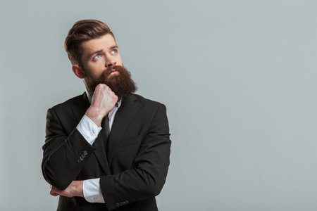 Thoughtful young bearded businessman in classic suit is looking away, on a gray backgroundの写真素材