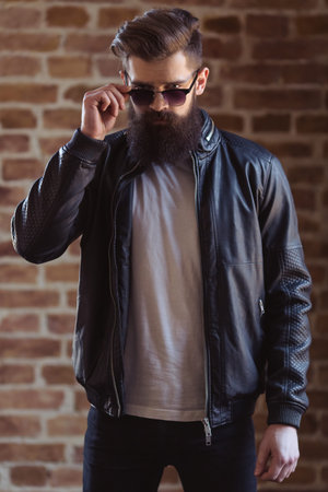 Stylish young bearded man in black leather jacket and sunglasses is looking at camera, standing against brick wallの写真素材
