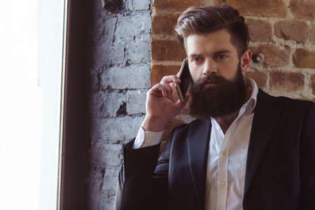 Handsome young bearded businessman in classic suit is talking on the mobile phone while standing near the windowの写真素材